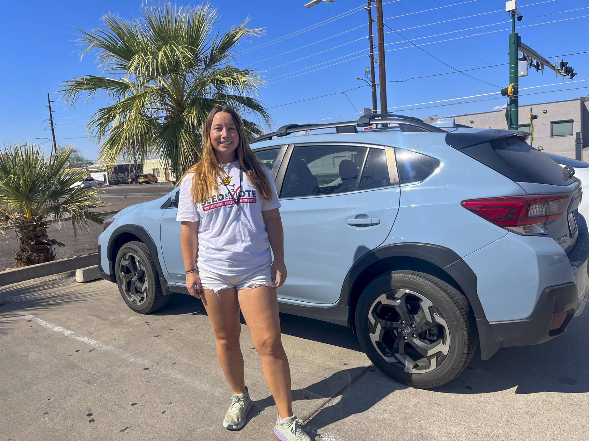 A Seed the Vote volunteer smiles in a sunny parking lot in Arizona, wearing a white Seed the Vote t-shirt and shorts, standing next to a light blue Subaru SUV with a palm tree and clear blue sky in the background.